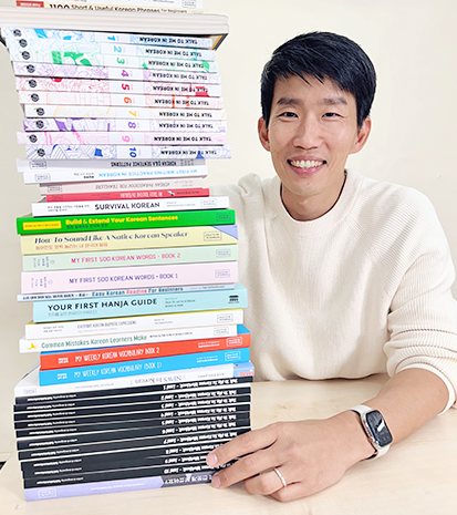 톡투미 인 코리안에서 출간한 한국어 학습 교재 44권과 선현우 대표 사진이다. 책 44권이 차곡차곡 쌓여있고, 그 옆에서 선현우 대표가 웃고 있다.