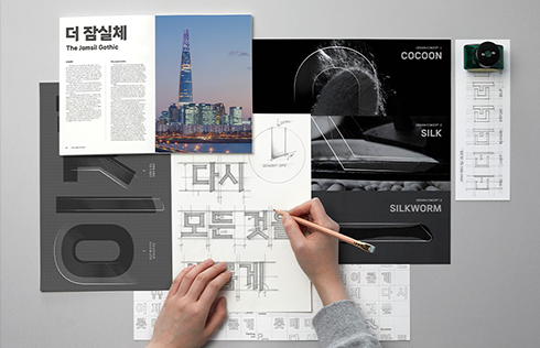 롯데마트에서 제작한 글꼴 ‘더잠실체’ 사진이다. 디자이너가 도면 위에 글자를 그리고 연구하는 모습을 담았다. 책상 주변에는 롯데월드타워 사진, 디자인 자료 등이 놓여 있다.