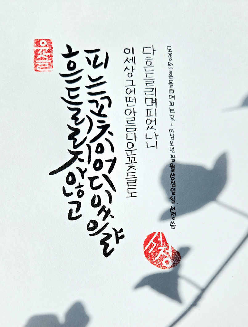 8월호 한 글 쓰기 참여작 사진이다. 하얀 종이 위에 검은색 붓으로 ‘흔들리지 않고 피는 꽃이 어디 있으랴 / 이 세상 그 어떤 아름다운 꽃들도 / 다 흔들리며 피었나니’라고 적혀있다. 배경에는 꽃의 그림자가 그려져 있다.