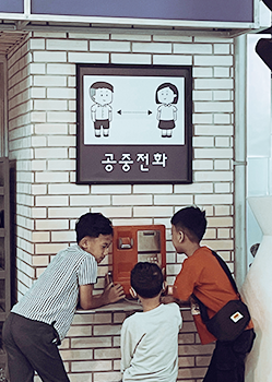 ‘2025년 내가 만난 한글 사진‧영상 공모’ 사진 분야 대상 수상작 ‘공중전화와 아이들’ 사진이다. 흰색 벽돌 벽에 붙은 ‘공중전화’ 안내판과 실제 공중전화기가 보이며, 안내판에는 남녀 아이 그림이 마주 보고 있는 픽토그램이 그려져 있다. 공중전화 앞에는 초등학생 정도로 보이는 남자아이 세 명이 모여 있는데, 한 아이는 고개를 숙이고 수화기를 들고 통화하는 듯하고, 다른 두 아이는 공중전화기를 흥미롭게 들여다보고 있다. 