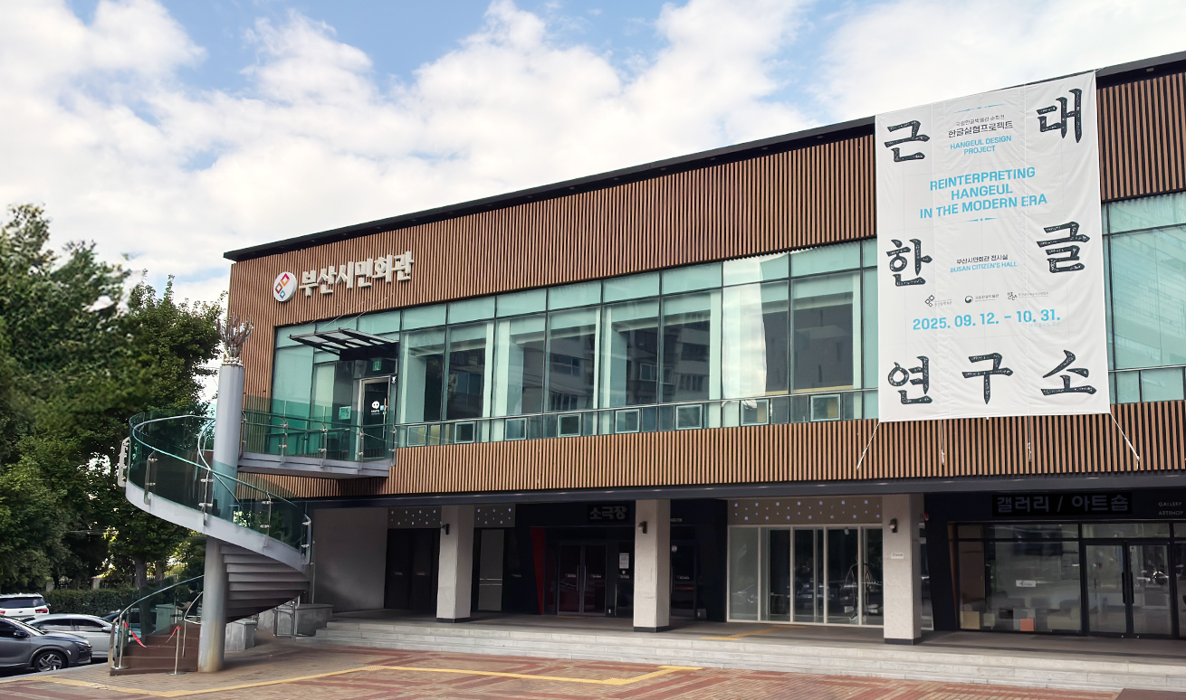 ‘근대한글연구소’ 전시가 진행 중인 부산시민회관 전경 사진이다. 건물 외벽은 수직으로 된 갈색 목재 패널과 유리창으로 이루어져 있는 2층 건물이다. 건물 상단 왼쪽에는 ‘근대 한글 연구소’ 전시를 알리는 대형 현수막이 걸려있다.