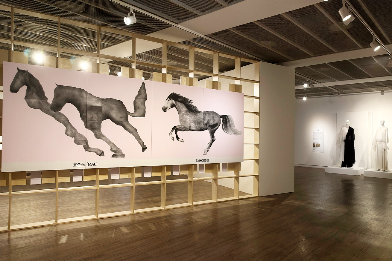 ‘근대한글연구소’ 부산 순회전 전시장 전경 사진이다. 나무 프레임 선반 중앙에 긴 직사각형 캔버스 두 장이 걸려있고, 말이 달리는 모습을 흑백으로 그린 대형 그림 두 점이 나란히 전시되어 있다.
