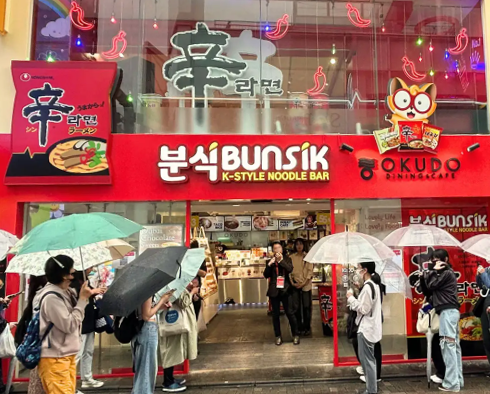 일본 하라주쿠에 열린 ‘신라면 분식’ 팝업스토어 사진이다. 빨간색 간판에 한글로 ‘분식’이라고 크게 적혀있으며, 신라면 봉지와 농심 너구리 캐릭터가 크게 배치되어 있다. 팝업 스토어 앞에는 사람들이 우산을 쓰고 입장을 기다리고 있다.