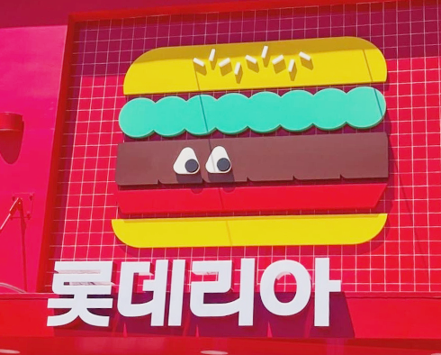 붉은색 배경에 큰 햄버거 일러스트가 그려져 있고, 햄버거는 노란 빵과 갈색 패티, 초록색 야채 층이 단순화된 도형으로 표현되어 있다. 햄버거 아래에는 흰색으로 ‘롯데리아’라는 글씨가 굵게 쓰여 있다.