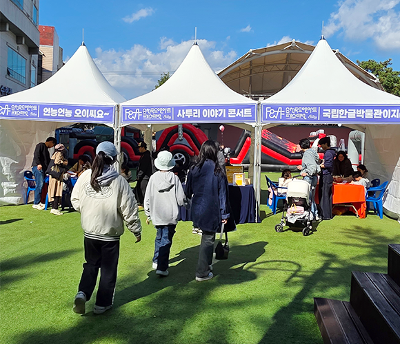 순천에서 열린 ‘찾아가는 사투리 이야기 콘서트’ 현장 사진이다. 흰색 천막 세 개가 나란히 서 있고, 각 천막의 현수막에는 흰 글씨로 각각 ‘언능언능 오이씨요~’, ‘사투리 이야기 콘서트’, ‘국립한글박물관이지라’라고 적혀있다. 천막 주변에는 체험하러 온 관광객들이 모여 있다.