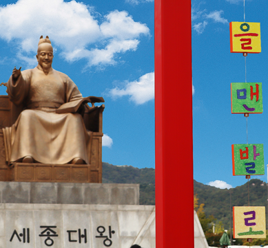 ‘2025 국립한글박물관 한글날 기념 문화행사’ 현장 사진이다. 푸른 하늘 아래 왼쪽에는 세종대왕 동상이 있다. 오른쪽에는 색색의 한글 조각들이 실에 매달려 공중에 떠 있다. ‘을’, ‘아’, ‘자’, ‘면’, ‘발’, ‘기’ 등 글자가 네모난 조각 위에 적혀있고, 각각 초록, 빨강, 노랑, 보라색 등 다양한 색으로 칠해져 있다.