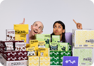 다양한 색깔과 디자인의 ‘한글과자’ 상자들이 쌓여있고, 상자에는 한글 글자 모양과 캐릭터 그림이 그려져 있다. 가운데에는 두 사람이 나란히 앉아 있는데, 왼쪽에 있는 타일러 라쉬는 웃는 표정으로 위를 가리키고 있고, 오른쪽에 있는 니디 아그르왈은 입술을 오므리며 고개를 살짝 젖히고 위를 가리키고 있다.