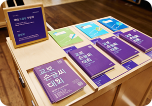 책상이 놓인 실내 공간 위에 다양한 색상의 책자들이 가지런히 전시되어 있다. 책자들은 초록색, 하늘색, 파란색, 보라색 등으로 구성되어 있으며, ‘교보 손글씨 대회’ 관련 문구가 표지에 큼직하게 적혀있다. 왼쪽에는 책자들에 대한 설명이 담긴 입간판이 세워져 있고, ‘역대 으뜸상 수상작’이라는 문구가 적혀있다.