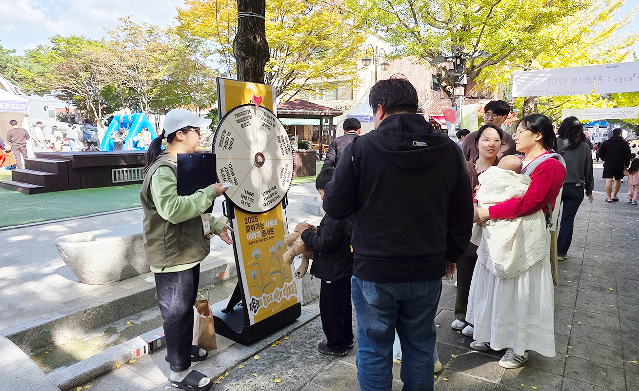 ‘찾아가는 사투리 이야기 콘서트’ 연계 체험 활동 사진이다. 야외 행사장에서 사람들이 체험 부스를 둘러보는 모습이다. 나무 아래에 돌림판 퀴즈가 있고, 사람들이 참여하고 있다.