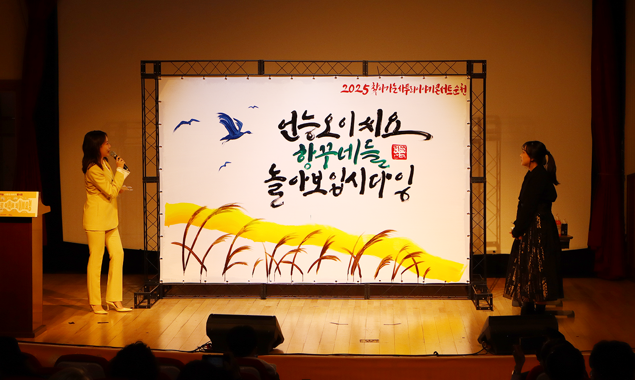 ‘찾아가는 사투리 이야기 콘서트’ 현장 사진이다. 무대 위 두 여성이 마이크를 들고 대화를 나누고 있다. 무대 중앙에는 갈대 그림과 함께 ‘언능 오이씨요 항꾸네들 놀아보입시다잉’이라고 손글씨로 적힌 커다란 그림이 있다.