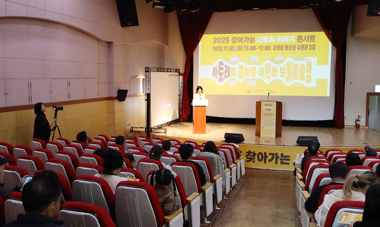 ‘찾아가는 사투리 이야기 콘서트’ 현장 사진이다. 강연이 진행되는 공연장 내부로, 객석에는 사람들이 앉아 있고 무대 앞 스크린에는 노란 배경의 ‘찾아가는 사투리 이야기 콘서트’ 화면이 비치고 있다.