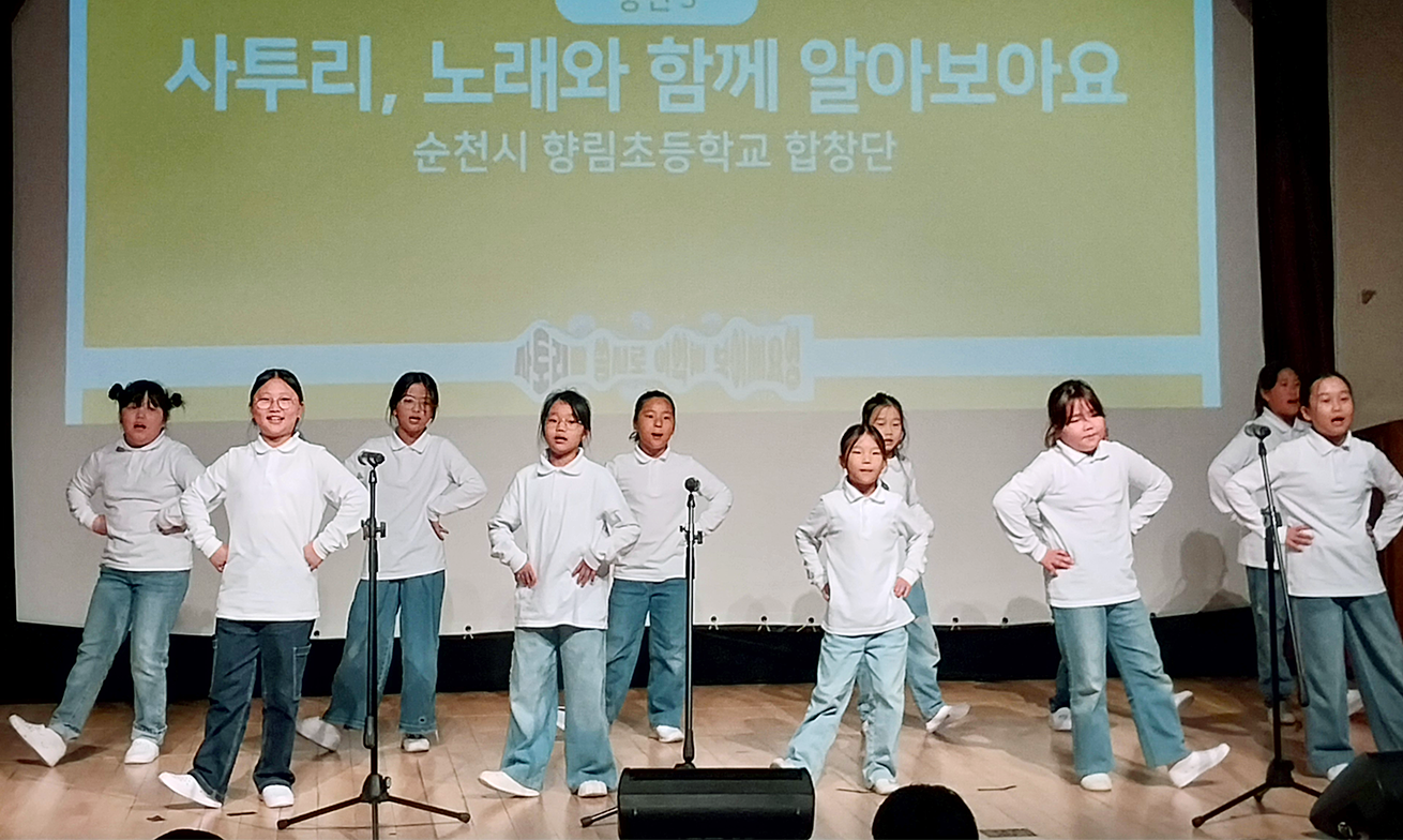 ‘찾아가는 사투리 이야기 콘서트’ 현장 사진이다. 흰 셔츠와 청바지를 입은 여덟 명의 학생이 무대 위에 서서 노래하며 율동하고 있다.
