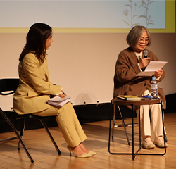 조정 시인 사진이다. 사회자는 노란 정장을 입고 노트를 손에 들고 있다. 조정 시인은 회색빛 단발머리에 갈색 코트를 입고 이야기하고 있다.