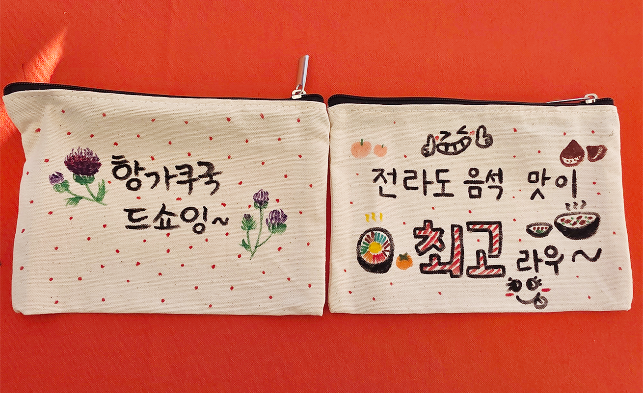 ‘찾아가는 사투리 이야기 콘서트’ 연계 체험 활동 사진이다. 천으로 된 필통 두 개가 펼쳐져 있고, 왼쪽에는 ‘항가쿠국 드쇼잉~’, 오른쪽에는 ‘전라도 음석 맛이 최고라우~’라는 문구가 손글씨로 적혀있다. 필통에는 보라색 엉겅퀴꽃과 하트, 별, 비빔밥 등 여러 모양이 그려져 있어 아기자기한 느낌을 준다.