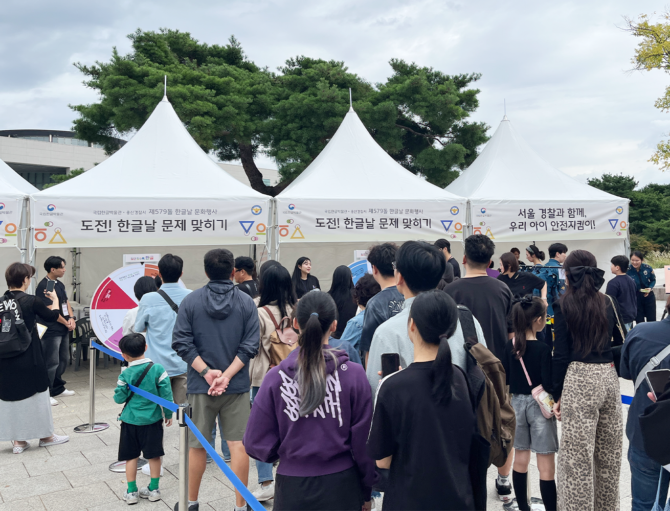 ‘2025 국립한글박물관 한글날 행사’ 현장 사진이다. 야외 행사장에서 여러 명의 사람이 하얀 천막 부스 앞에서 줄을 서 있다.