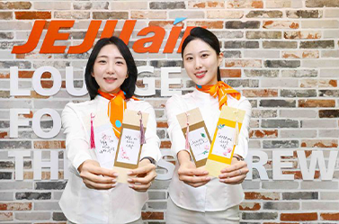 제주항공의 한글날 맞이 행사 현장이다. 두 명의 승무원이 흰색 제복에 주황색 스카프를 두르고 서 있으며, 손에는 한글 문구가 적힌 종이를 들고 있다.