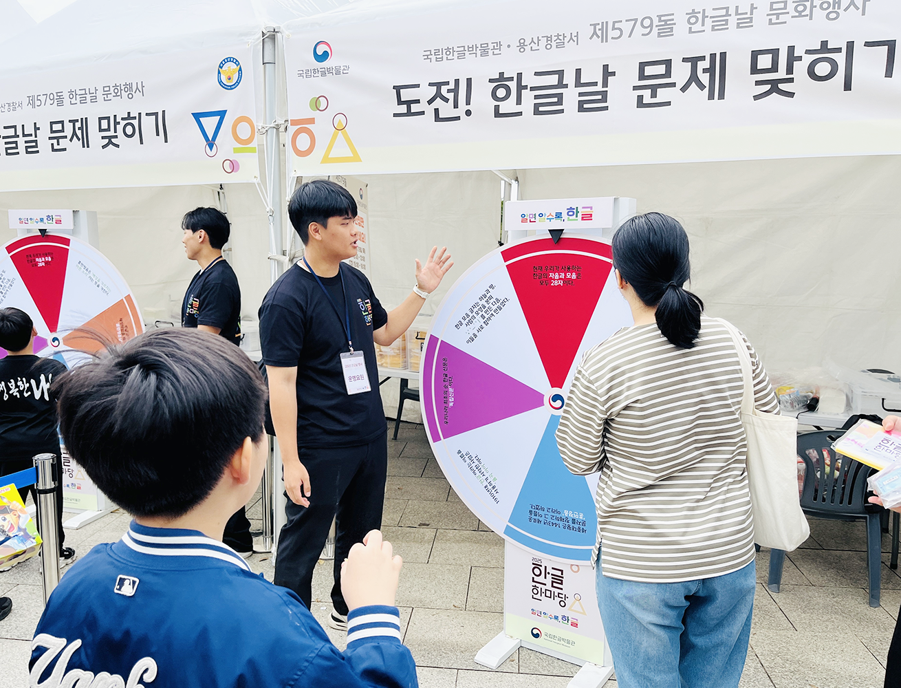 ‘2025 국립한글박물관 한글날 행사’ 현장 사진이다. 진행자가 커다란 돌림판 앞에서 사람들에게 설명하는 장면이다. 돌림판에는 색깔별로 다양한 퀴즈 항목이 적혀있고, 그 앞에는 어린이와 학부모가 모여 집중하고 있다.