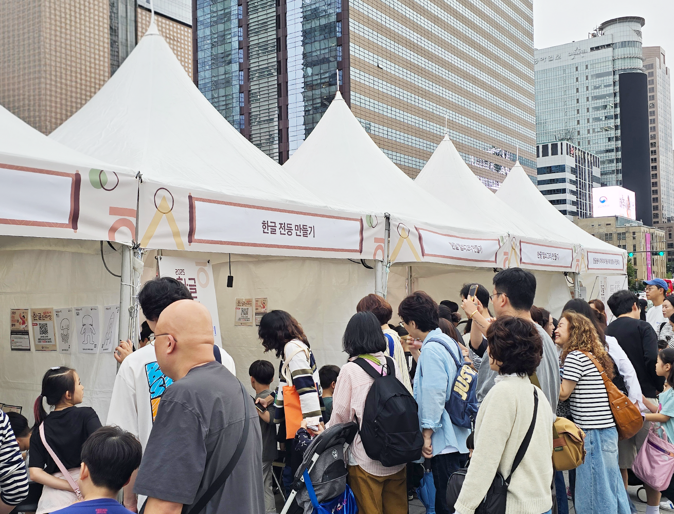 ‘2025 한글 한마당’ 국립한글박물관 체험 행사 현장 사진이다. 도심 속 야외 행사장에 설치된 하얀 천막 부스들이 줄지어 서 있고, 사람들로 북적이는 모습이다.