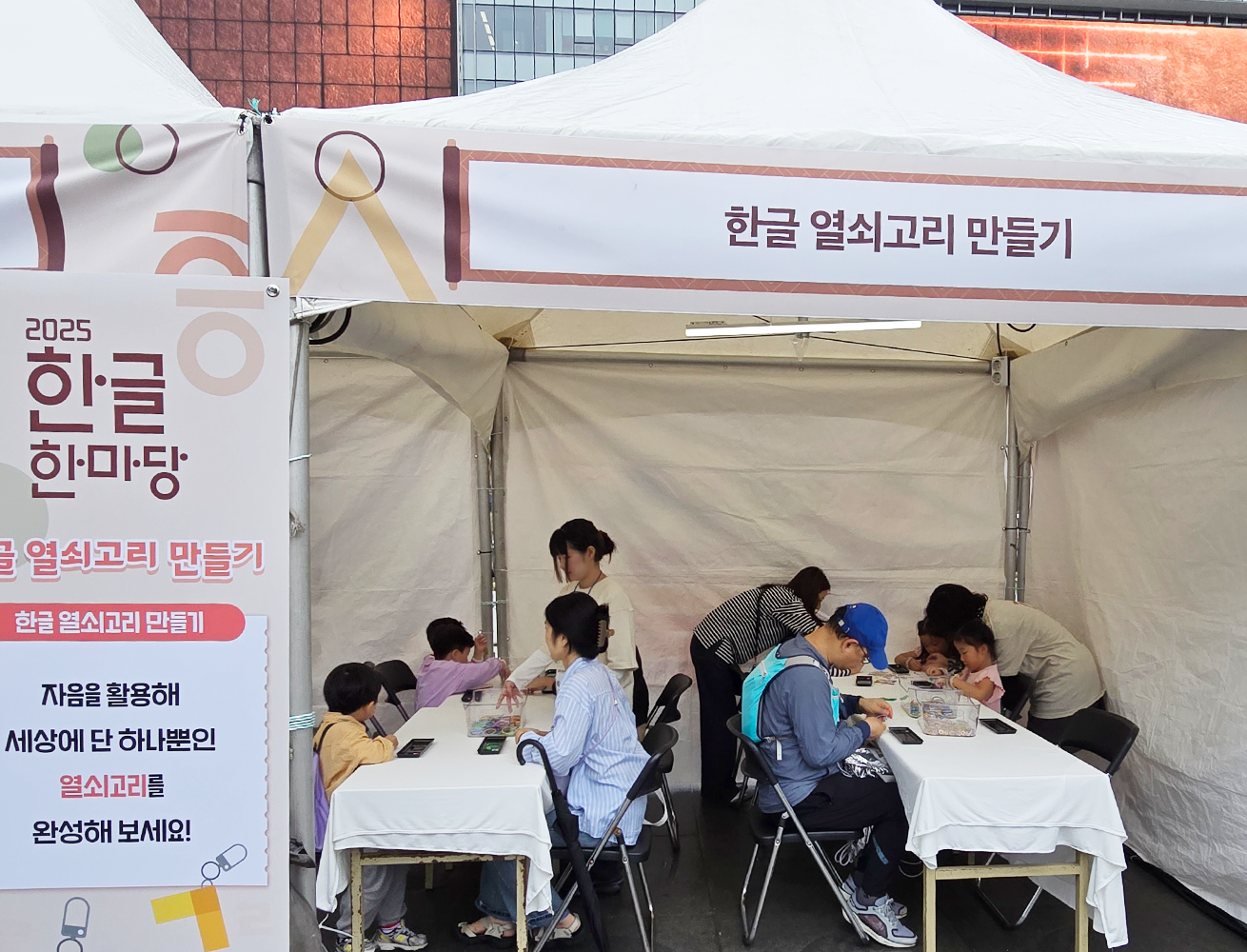‘2025 한글 한마당’ 국립한글박물관 체험 행사 현장 사진이다. ‘한글 열쇠고리 만들기’ 체험 부스 내부 장면으로, 테이블에 앉은 아이들과 학부모들이 함께 열쇠고리를 만들고 있다.