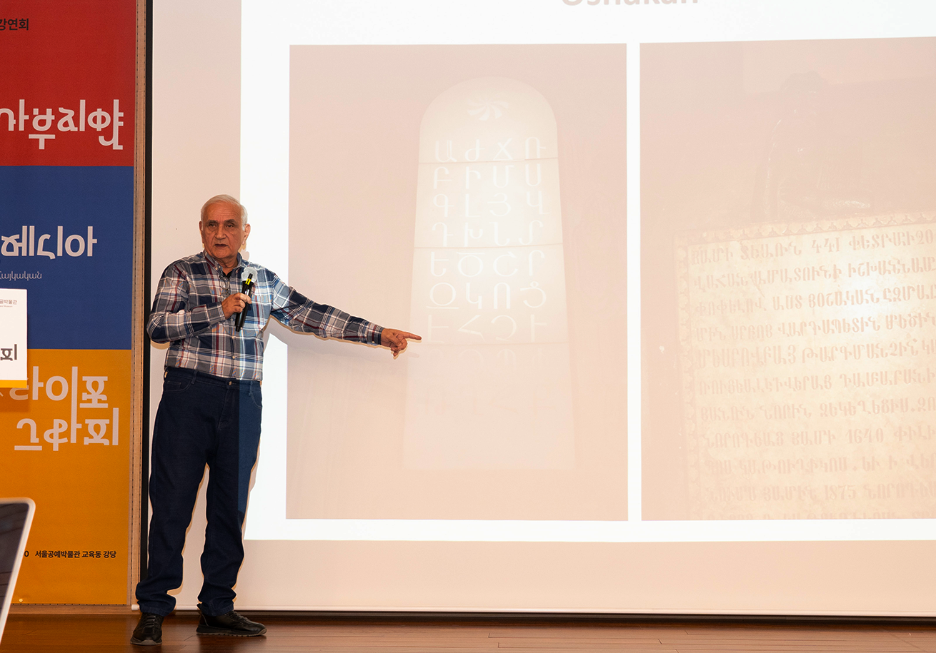 에딕 가부지얀의 강연 사진이다. 에딕 가부지얀이 손으로 스크린 속 아르메니아 유물을 가리키며 설명하고 있다.