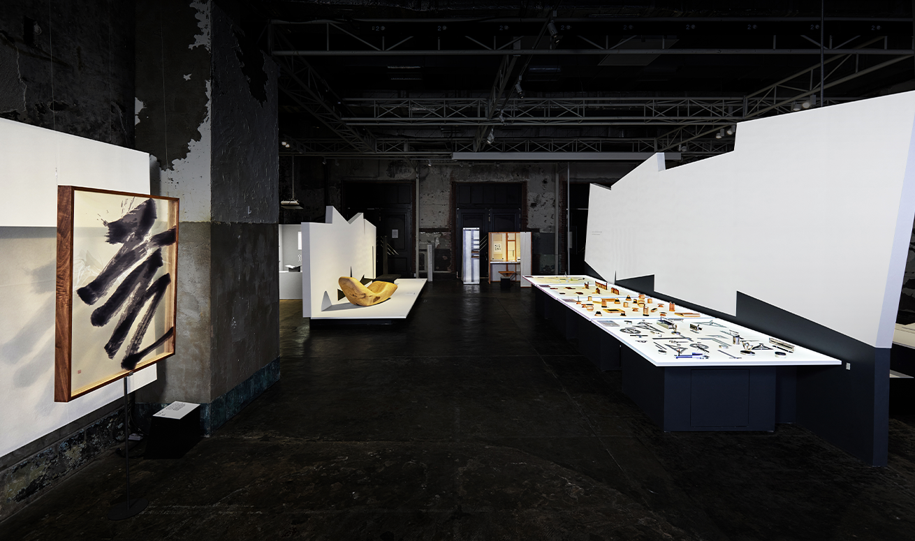 전시장 내부 사진이다. 흰색 종이 형태의 벽과 테이블이 배치되어 있다. 왼쪽 벽에 붓으로 쓴 대형 작품이 액자 형태로 걸려있고, 오른쪽에는 길게 뻗은 전시대 위에 다양한 작품이 배열되어 있다.