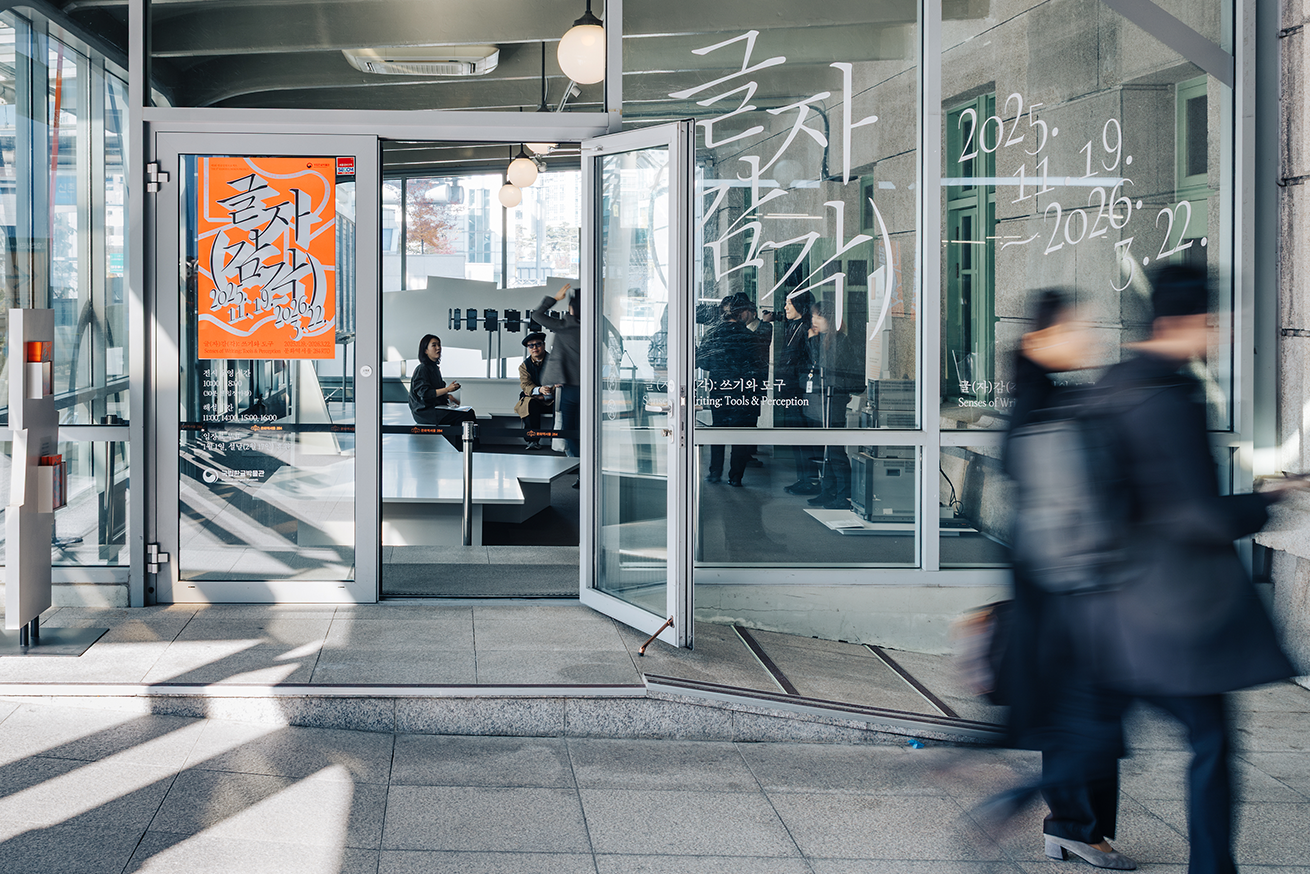 문화역서울 284 RTO 전시장 전경 사진이다. 유리문 너머로 사람들이 전시 개막을 준비하고 있으며, 왼편 유리문에는 주황색 배경에 흰 붓글씨로 쓰인 전시 포스터가 붙어 있다. 오른편 유리창에는 ‘글자 감각’이라는 전시명과 전시 기간이 큼직하게 붙어 있다.