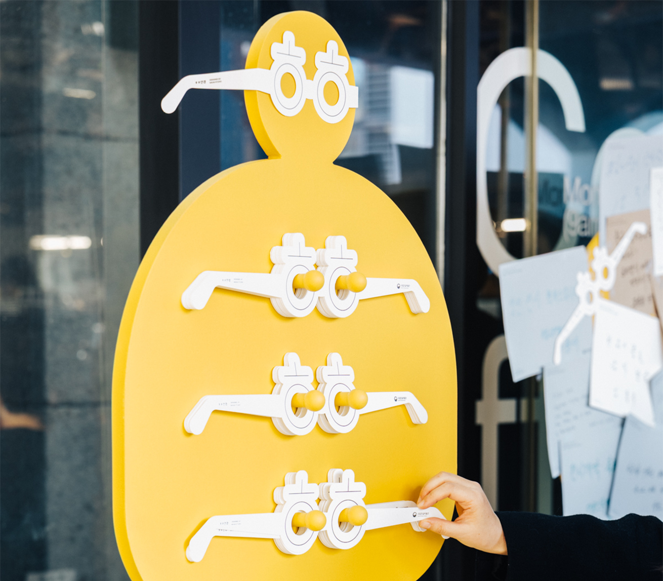 노란색 인체 모양의 보드가 세워져 있고, 그 보드에 ‘히읗’ 종이 안경이 여러 개 꽂혀 있다.