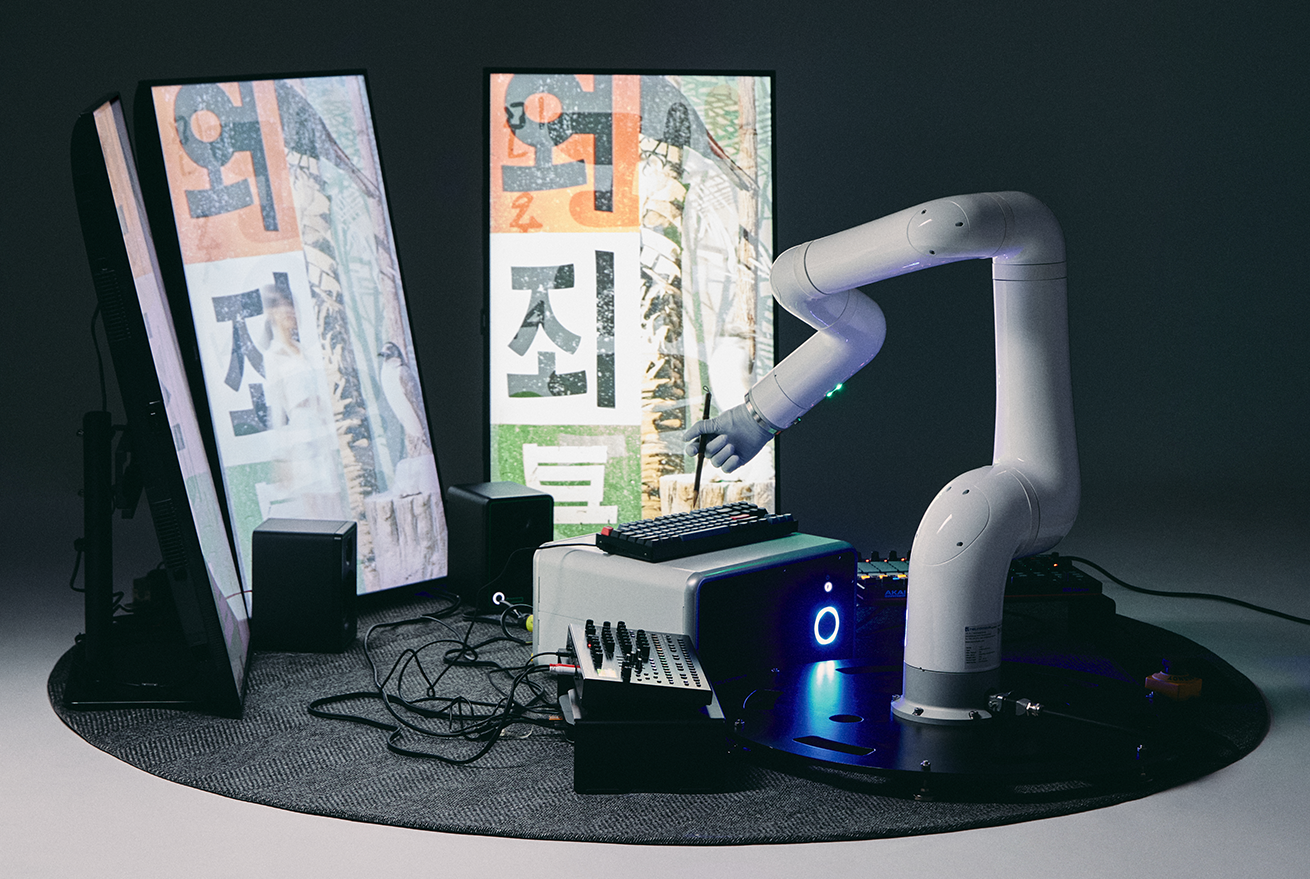 조영각의 ‘기획향’ 작품 사진이다. 로봇 팔 하나가 붓을 든 채 키보드 자판을 두드리고 있다. 그 앞에 있는 모니터에는 ‘죄’, ‘외’와 같은 글자와 추상적인 이미지가 그려져 있다.