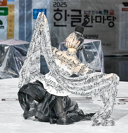 이상봉 디자이너의 ‘아리랑’ 작품이다. 한복의 족두리를 연상시키는 머리 장식을 한 모델이 아리랑 최초의 악보와 가사가 자수로 새겨진 옷을 입고 무대 위에서 포즈를 취하고 있다.