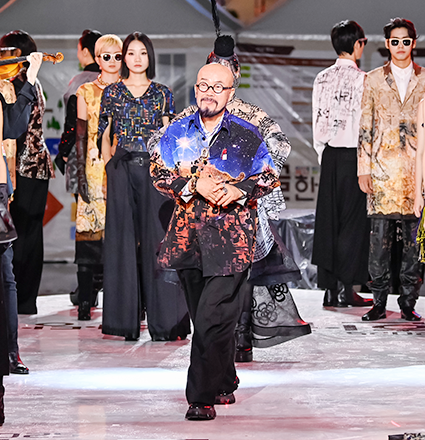 ‘2025 한글한마당(한글문화산업전)’에서 한글 패션쇼를 선보인 이상봉 디자이너 사진이다. 그는 검정 바지에 화려한 색감의 붉은빛과 푸른빛이 섞인 상의를 입고 있으며, 상의에는 한글 자모가 문양처럼 새겨져 있다.