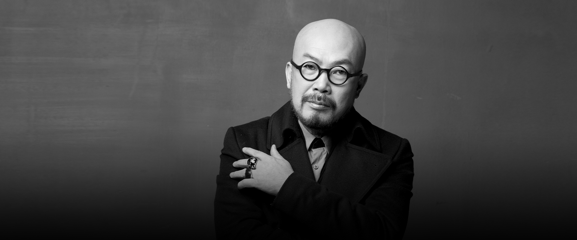 반갑습니다 사진. 이상봉 패션 디자이너의 모습이 담긴 흑백 사진이다. 이상봉 디자이너가 팔짱을 낀 채 카메라를 정면으로 응시하고 있다. 왼쪽 하단에는 주황색 박스 안에 ‘반갑습니다’ 문구가 적혀있다. 그 아래에는 기사의 제목 ‘패션에 한글의 멋을 입히다 이상봉 디자이너’가 쓰여있다.