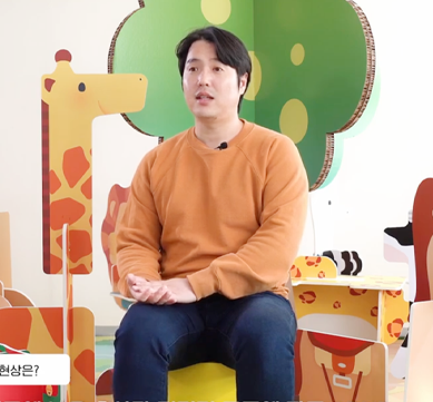 반갑습니다 사진. 주황색 긴팔을 입은 아토이 김형기 대표가 의자에 앉아 있다. 배경에는 아토이의 한글 글자 블록으로 만든 동물 모형이 있다.