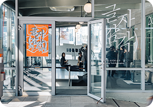 제5회 한글실험프로젝트 ‘글(자)감(각): 쓰기와 도구’ 전시 개막 현장 사진이다. 유리문 너머로 사람들이 전시 개막을 준비하고 있으며, 왼편 유리문에는 주황색 배경에 흰 붓글씨로 쓰인 전시 포스터가 붙어 있다. 오른편 유리창에는 ‘글자 감각’이라는 전시명과 전시 기간이 큼직하게 붙어 있다.