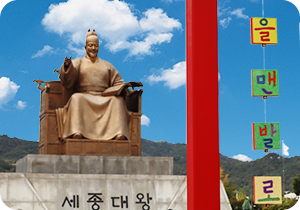 ‘2025 국립한글박물관 한글날 기념 문화행사’ 현장 사진이다. 푸른 하늘 아래 왼쪽에는 세종대왕 동상이 있다. 오른쪽에는 색색의 한글 조각들이 실에 매달려 공중에 떠 있다. ‘을’, ‘아’, ‘자’, ‘면’, ‘발’, ‘기’ 등 글자가 네모난 조각 위에 적혀있고, 각각 초록, 빨강, 노랑, 보라색 등 다양한 색으로 칠해져 있다. 