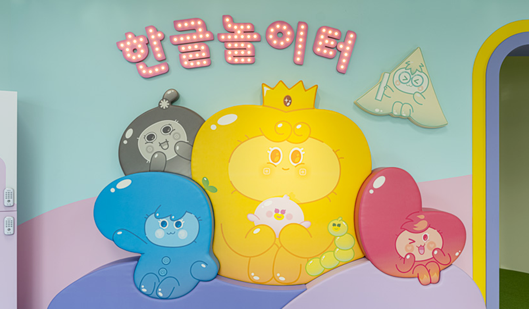 한글놀이터 입구 사진이다. 민트색 벽면에 '한글놀이터'라는 분홍색 간판이 걸려있고 그 아래로 노랑, 파랑, 분홍색의 귀여운 '말랑통통' 캐릭터들이 그려져 있다.