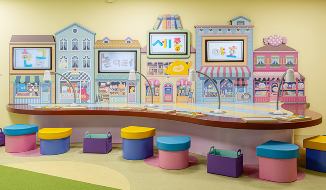 1부 '말랑통통 마을의 풍경'의 내부 공간 사진이다. 알록달록한 건물 모형이 늘어선 벽면 앞쪽으로 길쭉한 곡선 형태의 체험 책상과 알록달록한 의자들이 놓여있다. 마을 분위기로 꾸며진 체험 공간 곳곳에는 한글 자모를 활용한 놀이 요소들이 배치되어 있다.