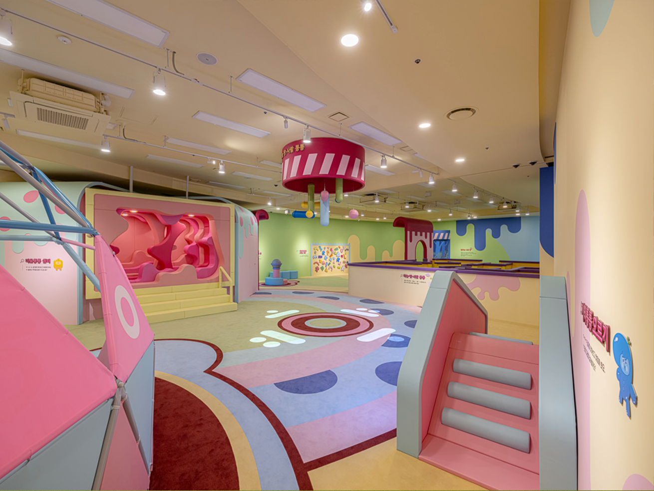 2부 ‘친구야! 한글놀이터에서 놀래?’ 내부 공간 사진이다. 전체적으로 분홍색과 하늘색 등 화사한 파스텔 톤이 사용되었으며, 바닥과 벽면은 부드러운 곡선과 아기자기한 무늬로 장식되어 있다.