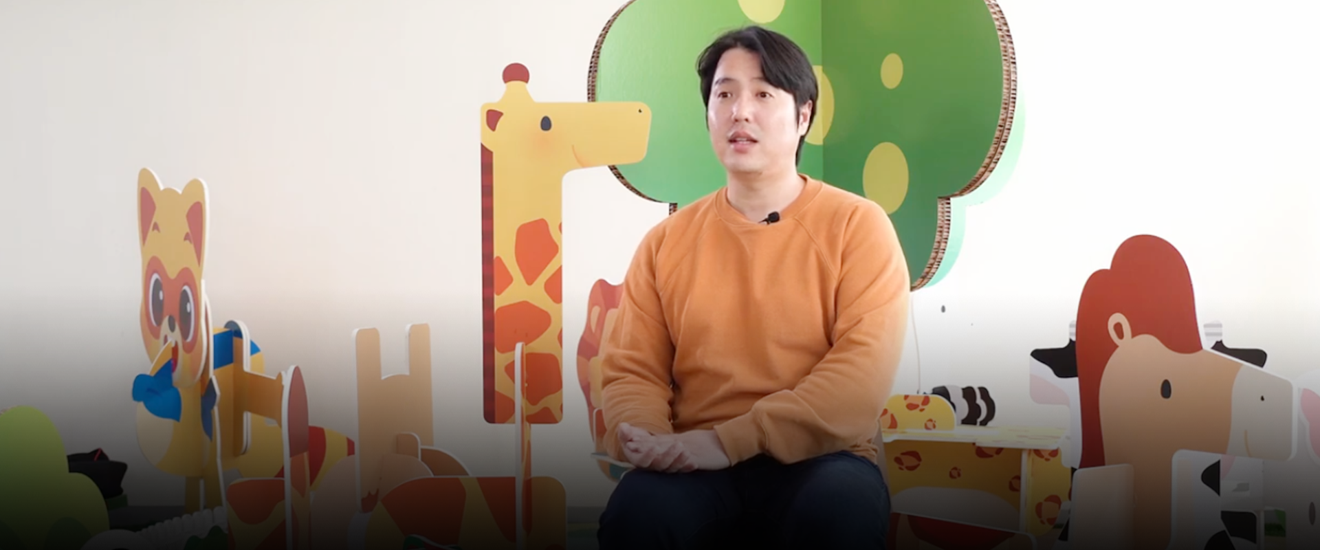 반갑습니다 사진. 주황색 긴팔을 입은 아토이 김형기 대표가 의자에 앉아 있다. 배경에는 아토이의 한글 글자 블록으로 만든 동물 모형이 있다. 왼쪽 하단에는 주황색 박스 안에 ‘반갑습니다’ 문구가 적혀있다. 그 아래에는 기사의 제목 ‘한글 블록으로 여는 배움의 즐거움, 아토이 김형기 대표’가 쓰여있다.