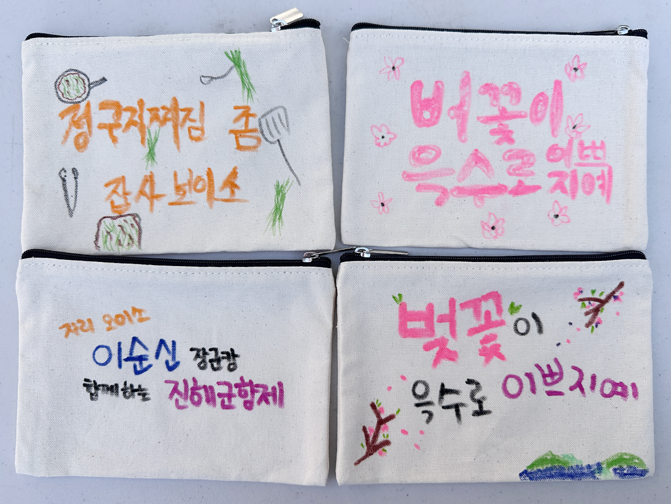 2025 사투리 체험 행사 ’파우치 만들기‘ 참여작이다. 천 재질의 파우치에 ’정구지찌짐 좀 잡사보이소‘, ’버꽃이 윽스로 예쁘지예‘, ’이순신 장군캄 함게하는 진해군항제‘ 등 손글씨와 간단한 그림이 알록달록한 색으로 그려져 있다.
