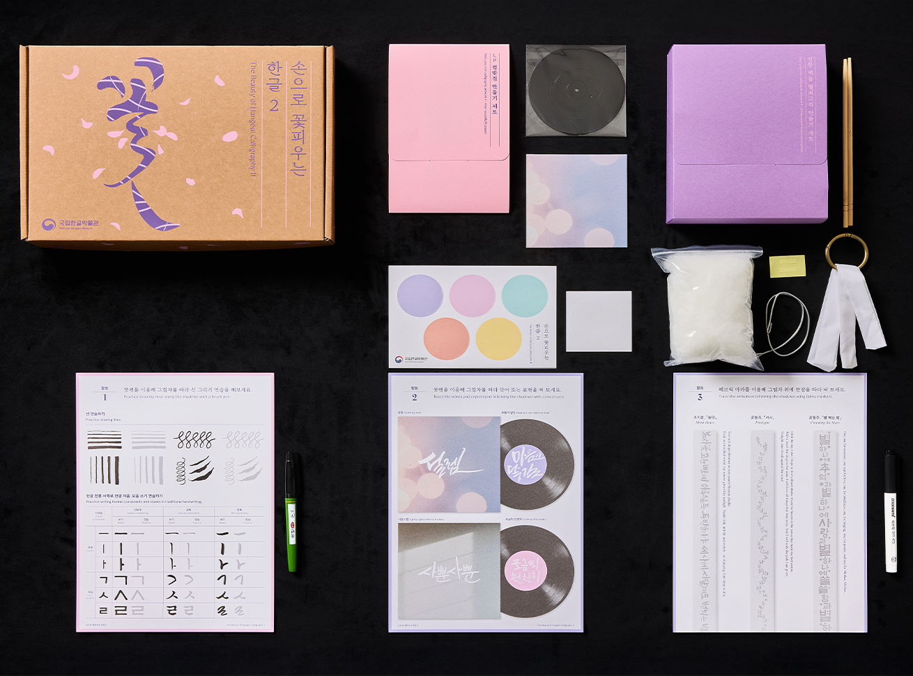 외국인 대상 교육_‘손으로 꽃피우는 한글’ 교구재 사진이다. 검은 배경 위에 한글 교육과 예술 활동을 위한 다양한 자료들이 가지런히 배열돼 있다. 왼쪽에는 분홍색과 보라색을 중심으로 한 교재 상자, 종이 자료 등이 정돈된 세트 형태로 놓여있다. 