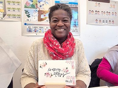 붉은 스카프를 두른 여성이 환하게 웃으며, 손 글씨로 작성한 ‘사이코지만 괜찮아’ 작품을 선보이고 있다.