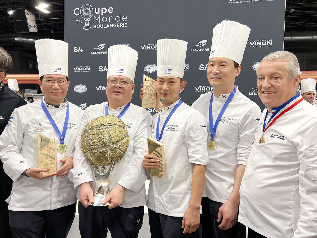 세계 제빵 월드컵 ‘2026 쿠프 뒤 몽드 드 라 불랑주리’에서 우승한 한국 국가대표팀 사진이다. 네 명의 한국 국가대표팀 제빵사들은 흰색 셰프 복장과 조리사 모자를 착용한 채 나란히 서 있으며, 손에는 상패와 트로피를 들고 있다. 그 옆에는 외국인 심사 위원이 함께 서 있다.