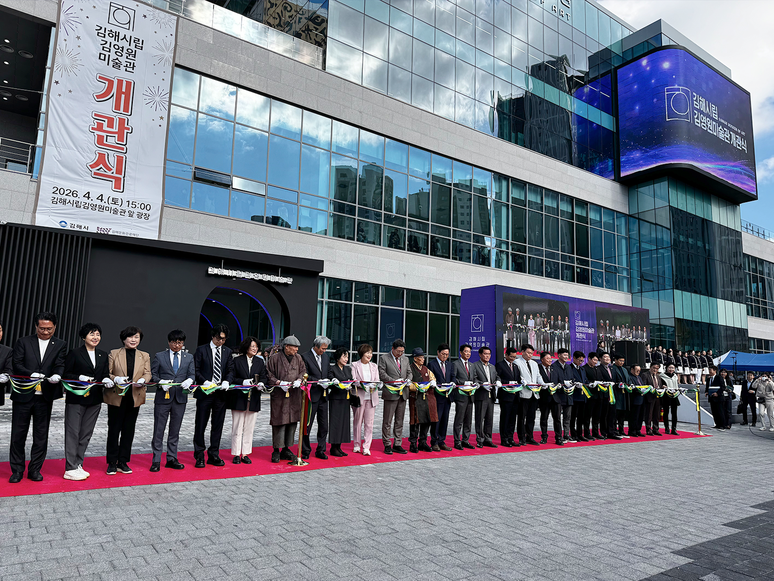김해시립김영원미술관 개관식 현장이다. 미술관 건물 앞에 관계자들이 줄지어 서 있고, 테이프를 가위로 자르는 커팅식을 개최하고 있다.