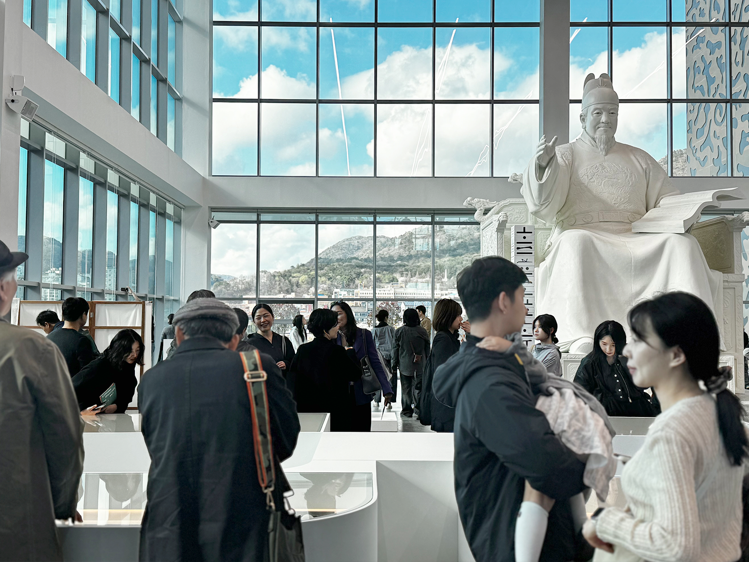 <글(자)감(각): 쓰기와 도구> 김해 순회전 현장 사진이다. 유리창으로 빛이 들어오는 공간에서 관람객들이 작품을 둘러보고, 뒤쪽에는 세종대왕 동상 원형이 있다.