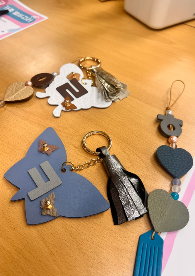 「한글 디자인 공방」 결과물 사진이다. 나비 모양의 키링에 한글 자음 ‘ㄹ’, ‘ㅋ’이 장식되어 있고, 테슬이나 나뭇잎, 하트 모양의 장식물이 달려있다.