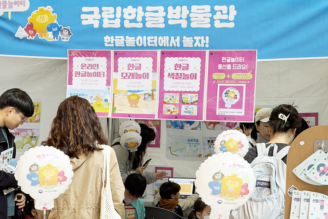 ‘한글놀이터에서 놀자!’ 행사 전경이다. 부스 상단에는 ‘한글놀이터에서 놀자!’라는 문구와 함께 다양한 체험 안내 포스터가 걸려 있다. 부스 앞에는 아이들이 앉아 활동에 참여하고, 주변에서는 어른들이 지켜보거나 함께 체험을 돕고 있다.