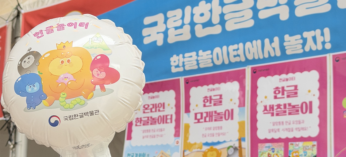 국립한글박물관 체험 부스를 알리는 홍보 배너와 풍선이 함께 보이는 사진이다. 왼쪽에는 ‘말랑통통 한글 요정’ 캐릭터가 그려진 둥근 풍선에 ‘한글놀이터’ 문구가 적혀있다. 오른쪽 배경에는 ‘한글놀이터에서 놀자!’라는 문구와 함께 온라인 한글놀이터, 한글 모래놀이, 한글 색칠놀이 안내 포스터가 나란히 걸려 있다.