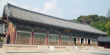 불국사 무설전 건물 전경을 담은 사진이다. 기와지붕을 얹은 길게 이어진 전통 한옥 건물이 정면으로 보이며, 기둥과 문이 일정한 간격으로 배치되어 있다. 건물 앞에는 돌로 된 기단과 계단이 놓여 있고, 뒤쪽에는 나무와 산이 어우러진 자연 배경이 펼쳐져 있다.