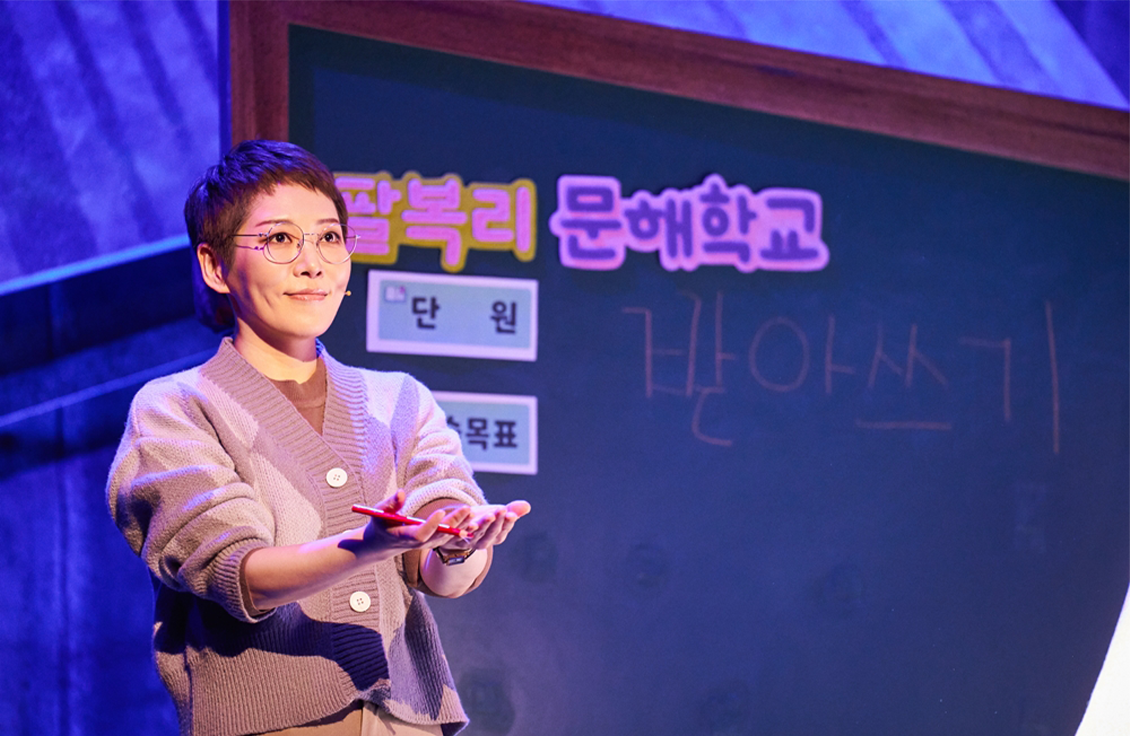 뮤지컬 ‘오지게 재밌는 가시나들’ 공연 장면이다. 팔복리 문해학교의 한글 선생님이 두 손을 내밀며 학생들을 격려하고 있다. 뒤편 칠판에는 ‘팔복리 문해학교’와 ‘단원’이라는 글자가 적혀있다.