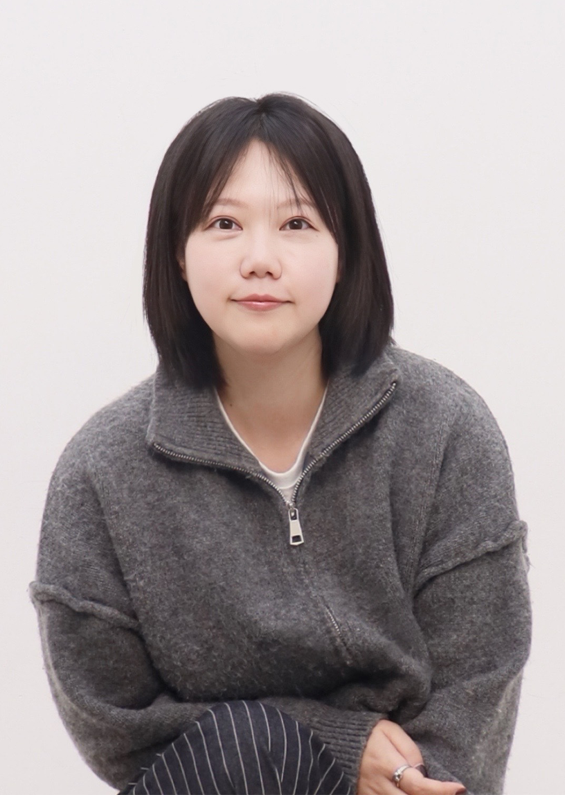 김하진 작가 사진이다. 회색 상의를 입은 김하진 작가가 카메라를 바라보며 미소를 짓고 있다.